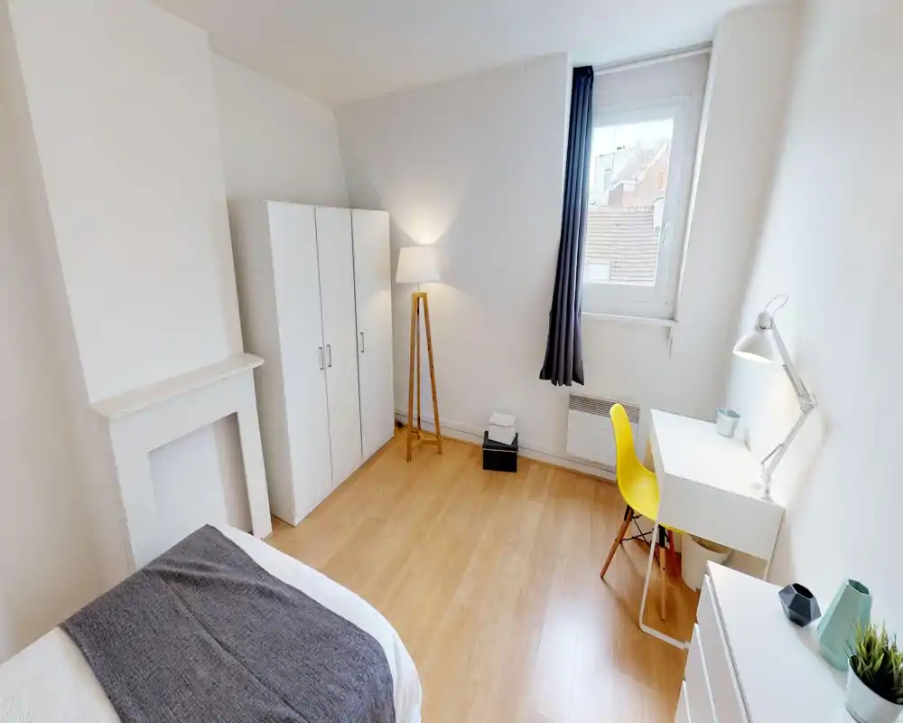 Chambre lumineuse et minimaliste avec un lit visible au premier plan, une armoire blanche, un sol en bois et un petit bureau près de la fenêtre — idéale pour une personne seule ou un couple.