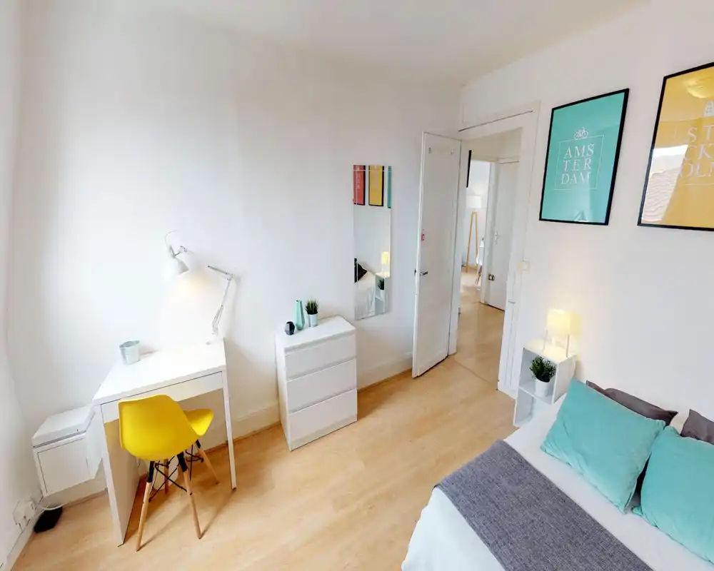 Chambre claire et soignée avec un lit confortable, coussins turquoise et lampe de chevet ; un petit bureau avec chaise jaune offre un coin travail.
