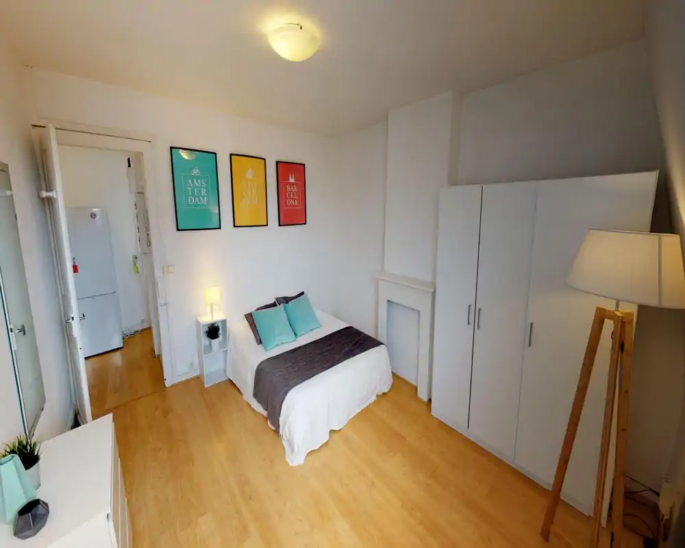 Chambre lumineuse et soignée avec un lit double en linge blanc et coussins turquoise, une lampe de chevet, une armoire et des affiches décoratives — idéale pour une annonce conviviale.