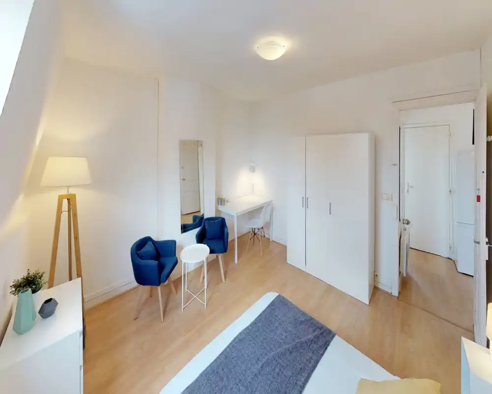 Bright, neatly styled bedroom with a visible double bed, two blue armchairs and a small desk. The room features light wooden floors, a large wardrobe and warm accent lighting for a cozy atmosphere.