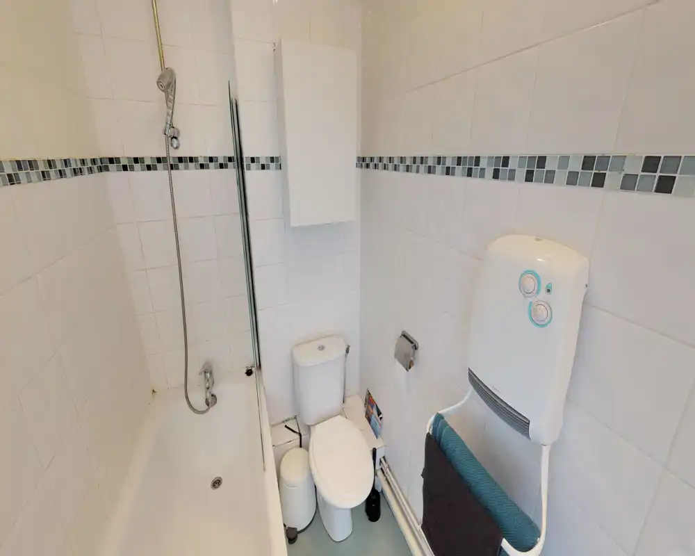 Compact white-tiled bathroom with a bath/shower combination, toilet, wall-mounted heater, and storage cabinet. Clean, bright and photographed from the doorway to show fixtures.