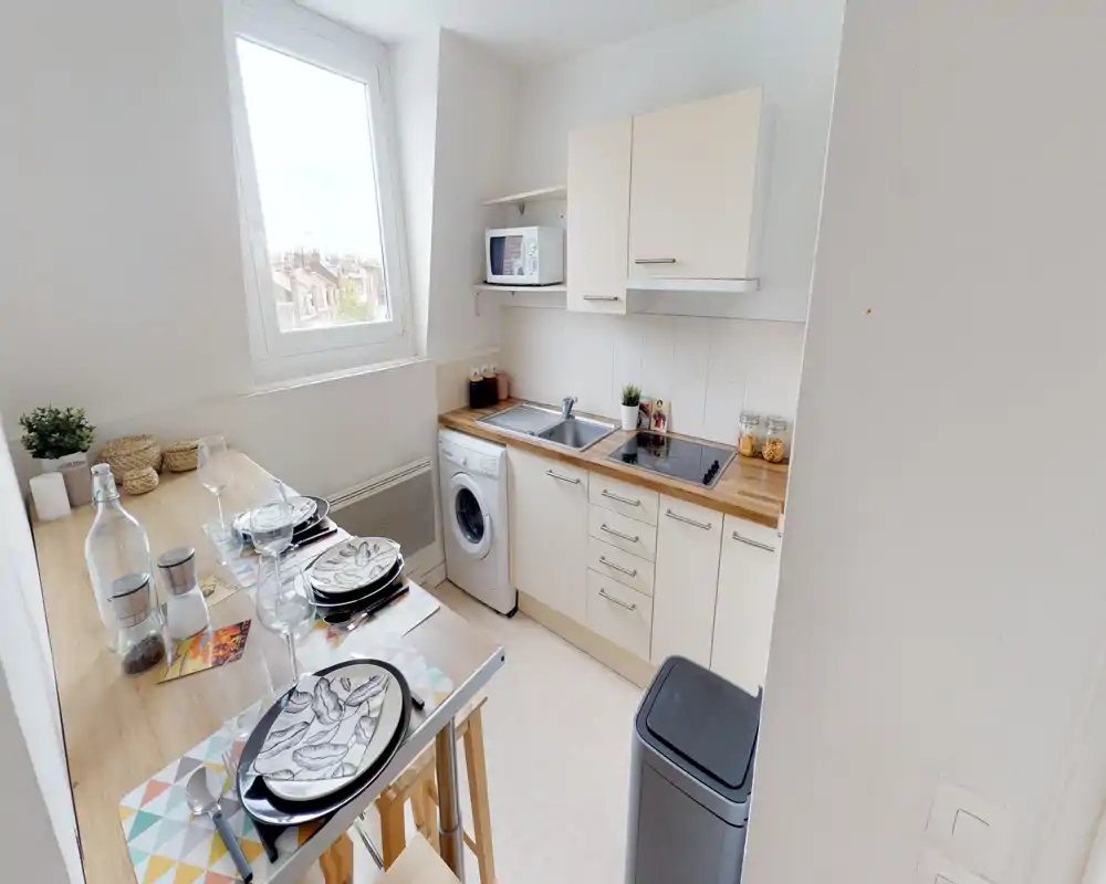 Compact, bright kitchenette with a two-burner hob, stainless sink, washing machine and a breakfast bar set for two. Natural light from the window and warm wood countertops create a welcoming small-space kitchen.