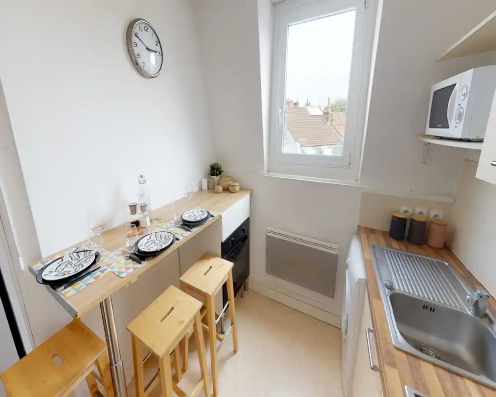 Compact fitted kitchen with wooden countertops, sink, microwave and a small breakfast bar with stools — bright window light and neat place settings.