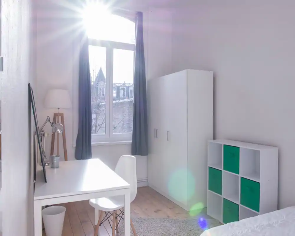 Coin chambre lumineux et minimaliste avec bureau, armoire et étagère; la lumière naturelle entre par une grande fenêtre et donne une impression d’air et de clarté.