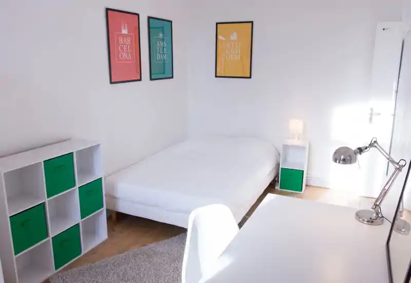 Chambre lumineuse et minimaliste avec un lit blanc, une petite table de chevet avec lampe, des cube de rangement verts et des affiches encadrées au mur.