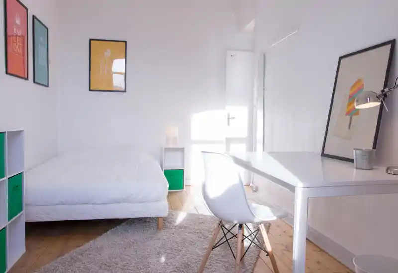 Chambre lumineuse et minimaliste avec lit bien fait, bureau blanc et chaise moderne. La lumière naturelle inonde la pièce et des affiches colorées apportent une touche vive — idéale pour une annonce montrant une chambre simple, propre et accueillante.