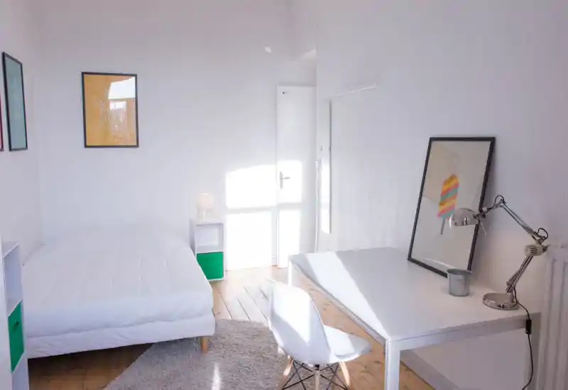 Chambre claire et minimaliste avec un lit double blanc, des étagères de chevet et un bureau près de la fenêtre. La lumière naturelle remplit la pièce et le tapis moelleux avec la lampe créent une atmosphère accueillante.