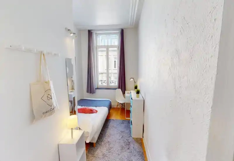 Chambre compacte et lumineuse avec un lit bien fait, tapis douillet et lumière naturelle provenant d'une grande fenêtre. Mobilier épuré de style scandinave et petit bureau pour un espace cosy et fonctionnel.