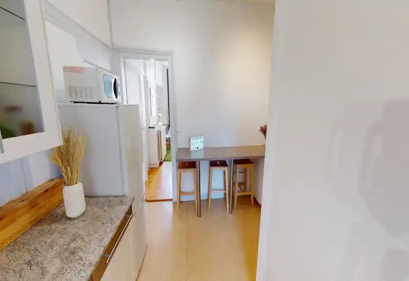 Coin cuisine compact avec un petit bar et trois tabourets en bois, plan de travail avec un vase décoratif et un réfrigérateur avec micro-ondes — idéal pour des repas rapides et un séjour convivial.