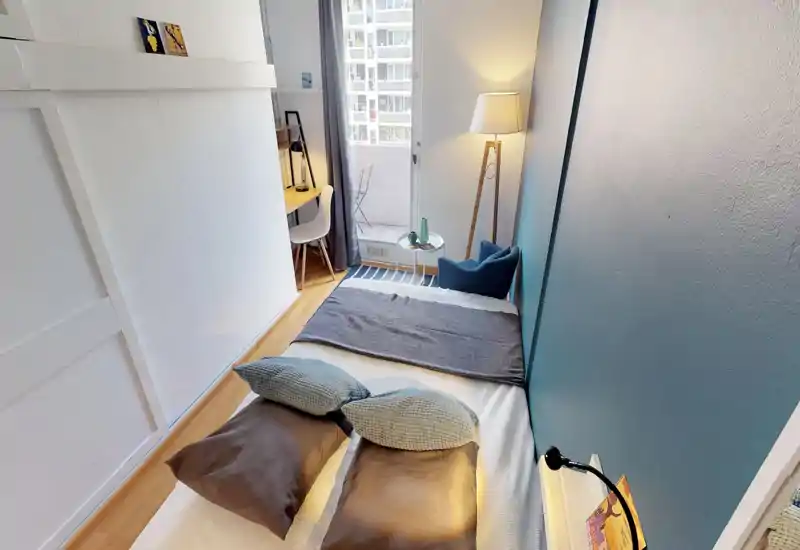 Bright, well-styled small bedroom with a double bed placed against a teal accent wall, soft pillows and a throw. A cozy corner with an armchair, floor lamp and a compact desk by the window opens onto a small balcony.