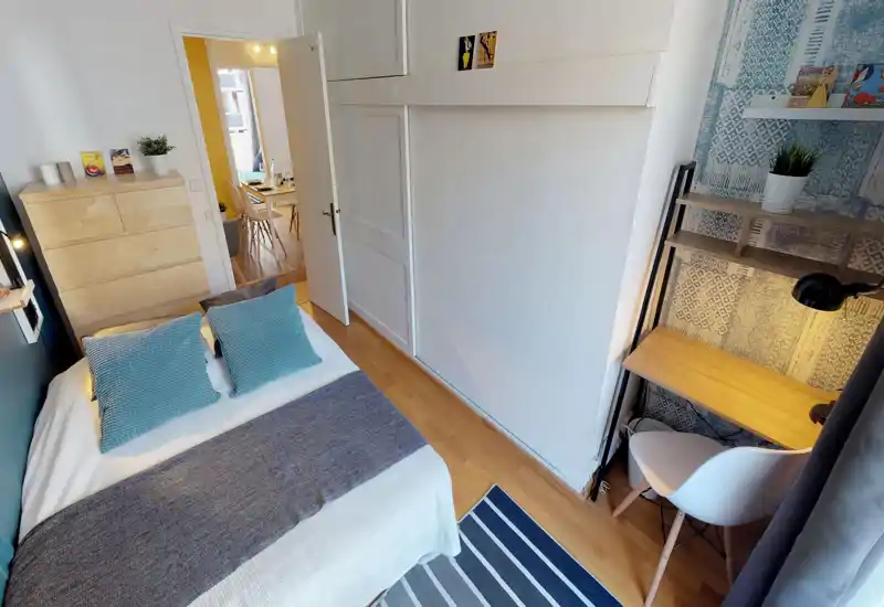 Bright, well-staged bedroom with a double bed dressed in white linens, teal accent pillows and a grey throw. A wooden chest of drawers, built-in wardrobe doors and a small workspace with shelving create a functional, cozy layout ideal for short stays.