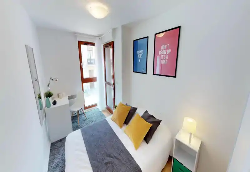 Camera da letto luminosa e ben allestita con letto matrimoniale, cuscini decorativi e una coperta accogliente; una scrivania e una sedia vicino alla finestra che lascia entrare luce naturale.