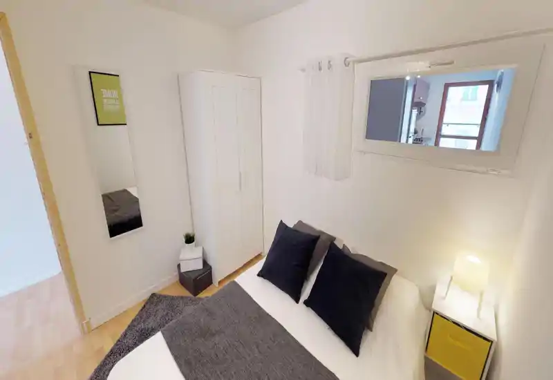 Compact, well-styled bedroom with a double bed, dark accent pillows, a white wardrobe and a bedside lamp providing warm light. Neutral walls and soft textiles create a calm, inviting atmosphere ideal for short-stay rentals.