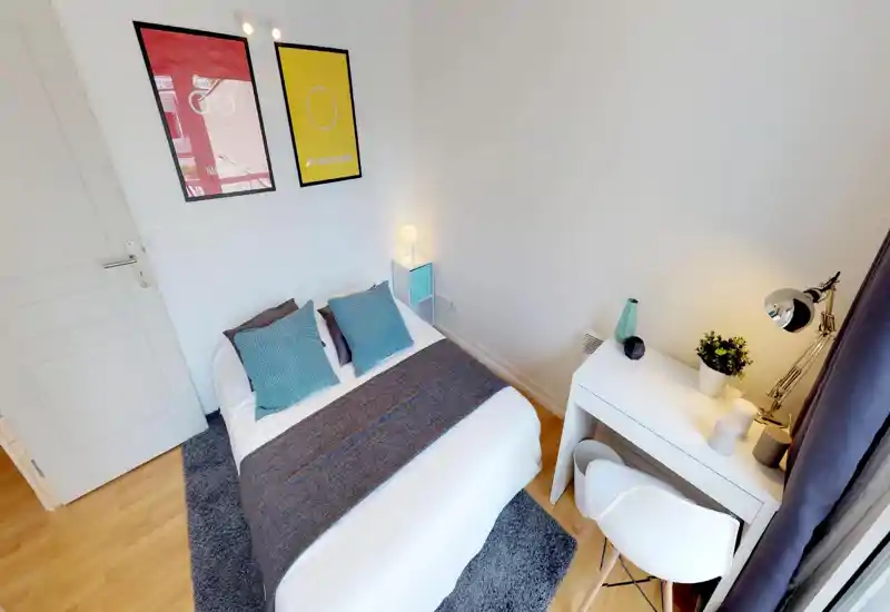 Bright, well-staged bedroom with a double bed, textured throw and two accent pillows; a small desk and lamp sit by the window creating a functional cozy corner.