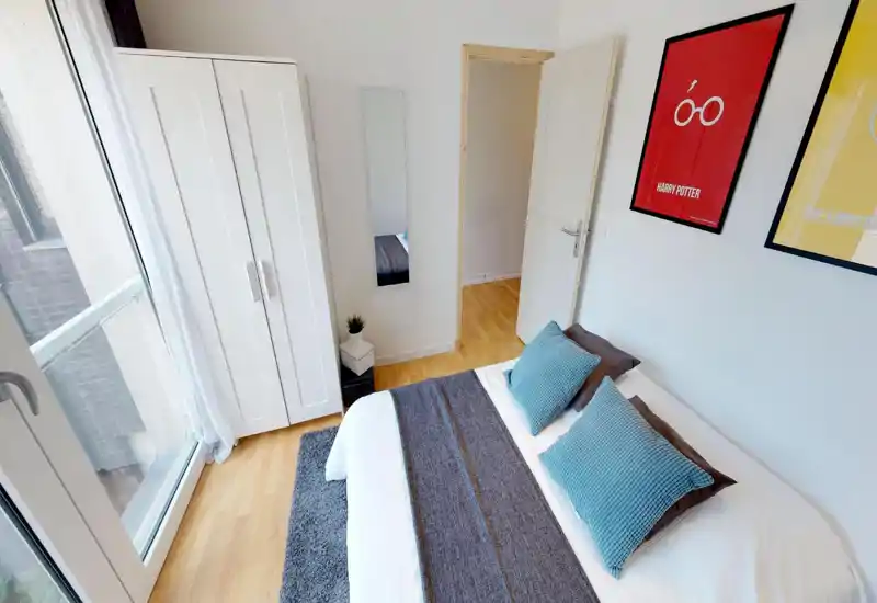 Bright, modern bedroom with a large window, white wardrobe and a neatly made double bed accented by blue cushions and framed wall art.