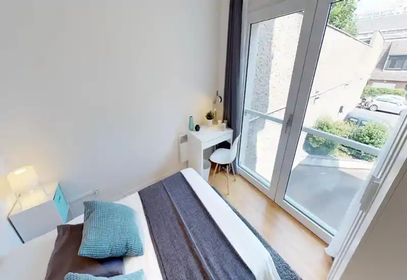 Bright, modern single bedroom with a neatly made bed, textured cushions and a small desk by a large floor-to-ceiling window letting in ample natural light.