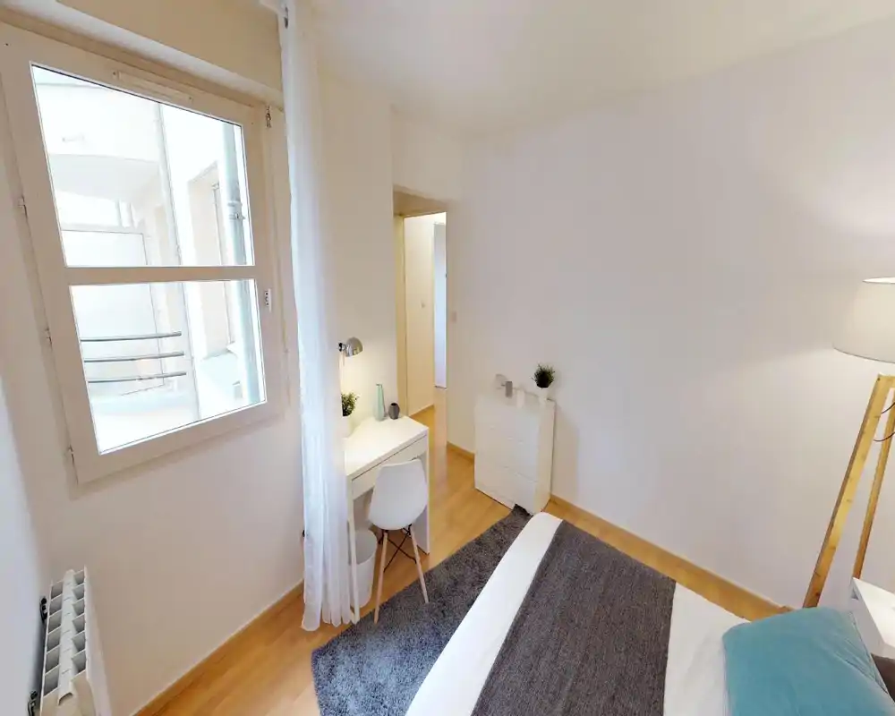 Bright, minimalist bedroom with a visible bed, bedside lamp, small desk and chest of drawers; neutral decor and wooden floor create a calm, welcoming atmosphere.
