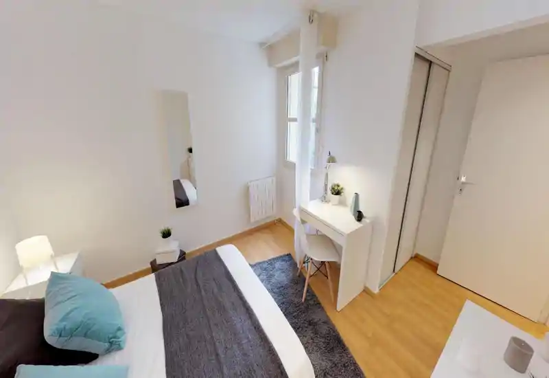 Bright, neatly styled bedroom with a large bed, bedside lamp, desk by the window and soft rug — modern minimalist decor ideal for a comfortable stay.