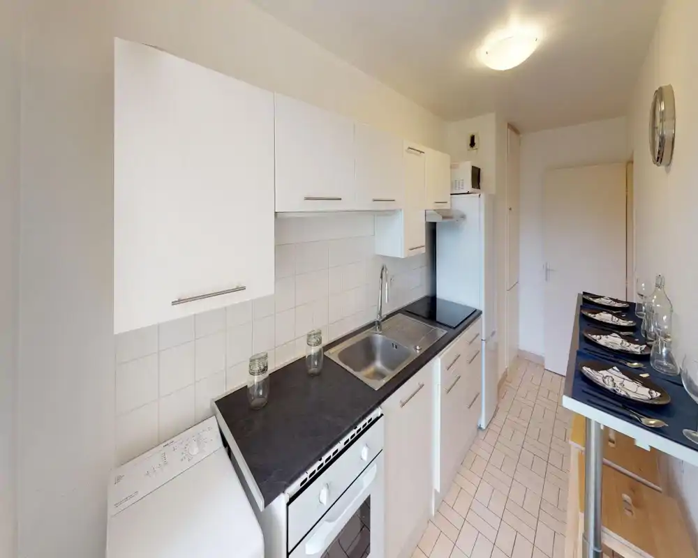 Cucina a corridoio compatta e moderna con mobili bianchi, piano scuro, forno integrato e lavello. Lo spazio è ordinato e ben illuminato, con un piccolo bancone apparecchiato per quattro persone.