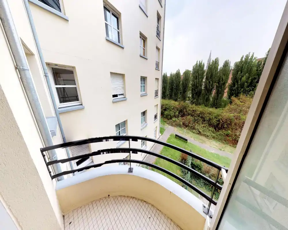 Piccolo balcone curvo che si affaccia su un cortile verde con alberi e un edificio adiacente; pavimento piastrellato e ringhiera in metallo visibili — perfetto per un caffè mattutino.