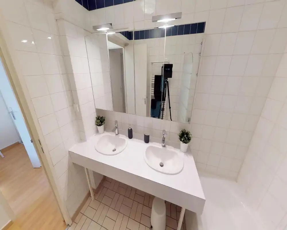 Bagno luminoso e pulito con doppi lavabi, grande specchio e piastrelle bianche. Buona illuminazione e arredo minimalista.