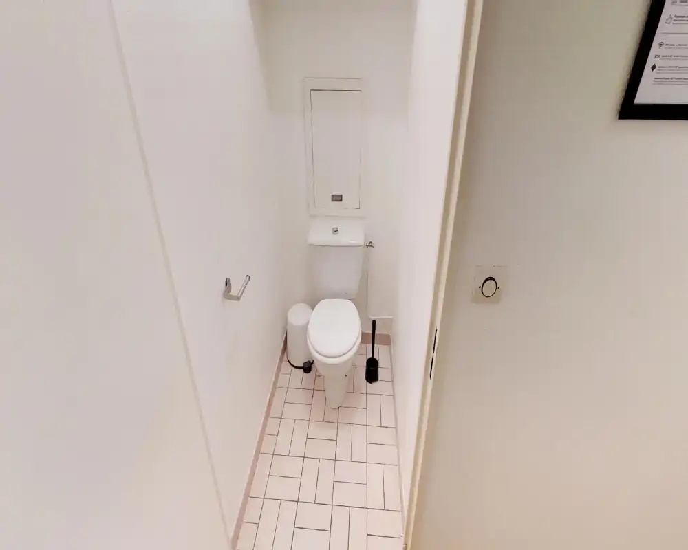 Piccolo WC pulito con wc bianco, scopino e piccolo cestino sulla pavimentazione piastrellata — luminoso e ben tenuto.