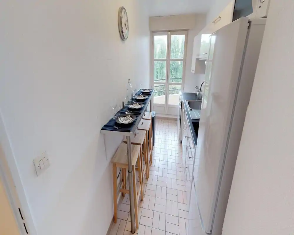 Cucina stretta e luminosa con bancone da pranzo per quattro, sgabelli, lavello, frigorifero e finestra/porta che lascia entrare luce naturale.