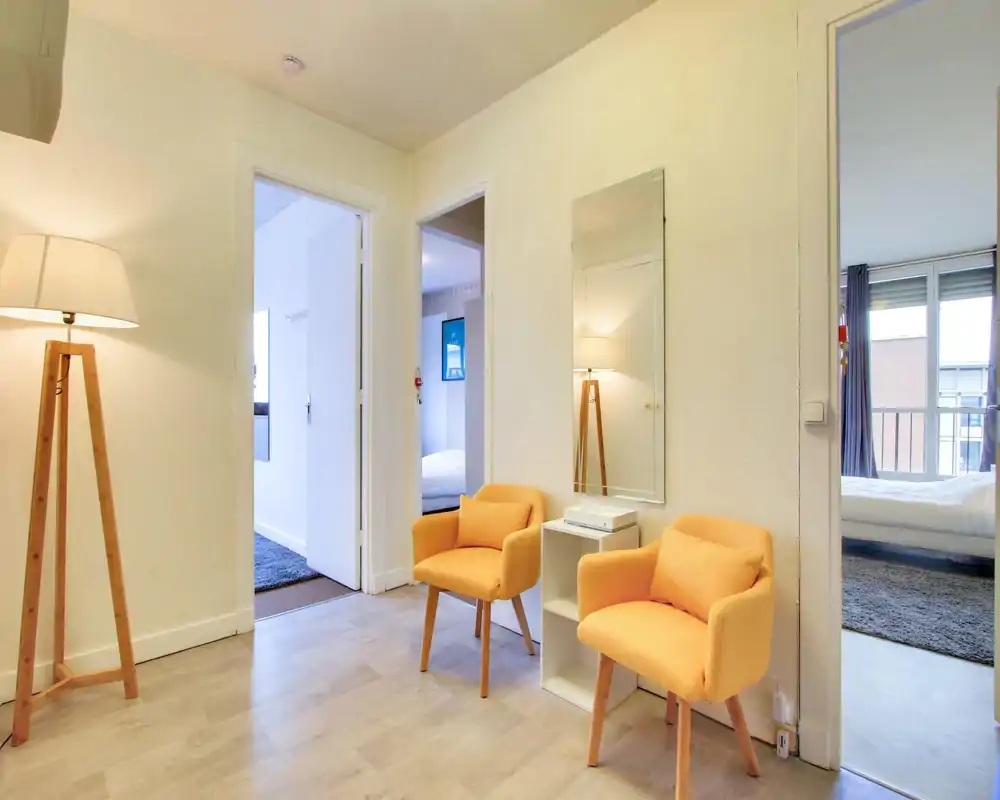 Bright, welcoming entrance/foyer with two mustard armchairs, a floor lamp and a wall mirror; neutral flooring and clear sightlines into adjacent bedrooms.