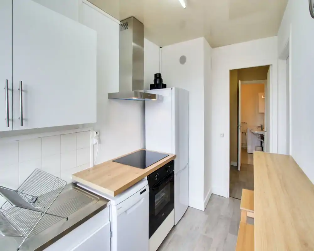 Bright, modern compact kitchen with white cabinets, wooden countertop, induction hob and integrated oven; practical layout with stools and a fridge.
