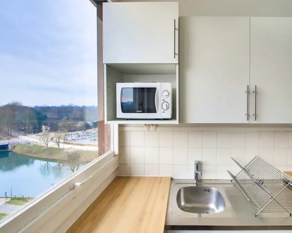 Bright compact kitchen with a stainless-steel sink, wooden countertop, white cabinets and a microwave tucked into a cupboard; large window offers a pleasant riverside view.
