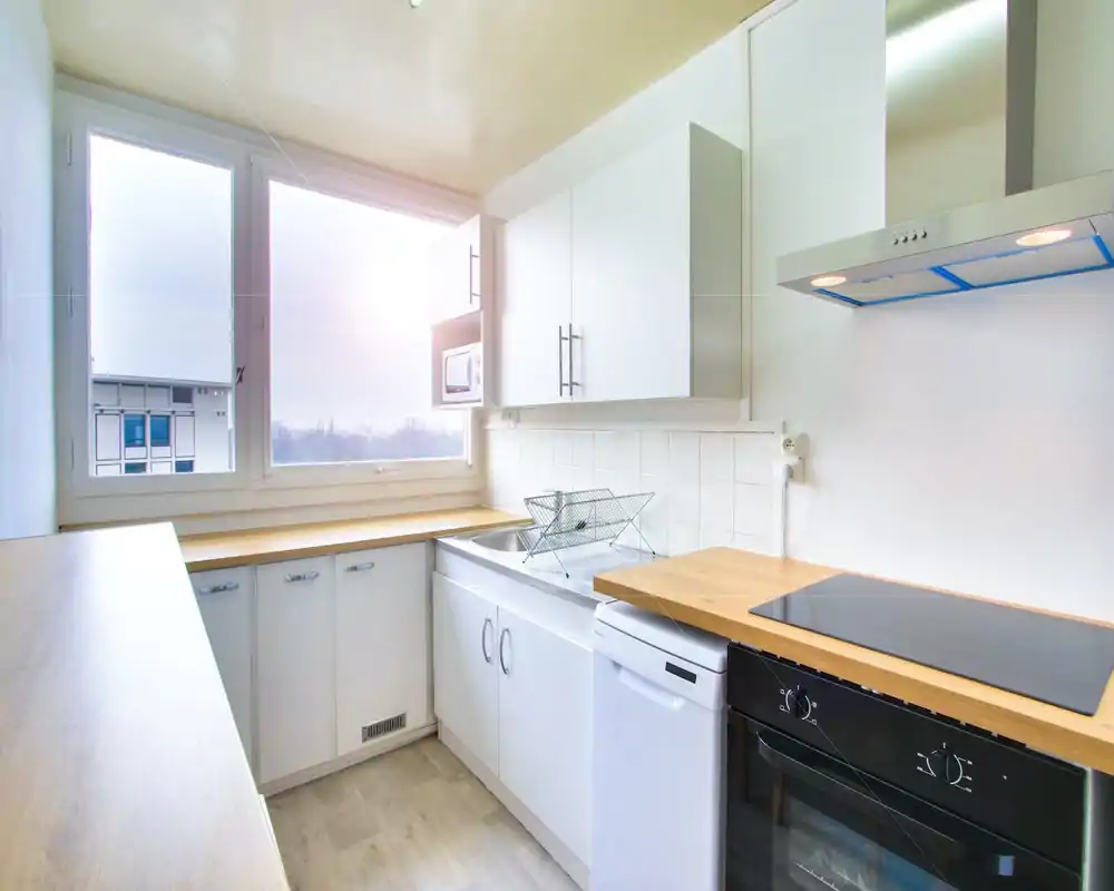 Bright compact kitchen with wooden countertops, white cabinets, and built-in appliances including an induction hob and dishwasher. Large window provides ample natural light and a clean, minimalist look ideal for a rental listing.