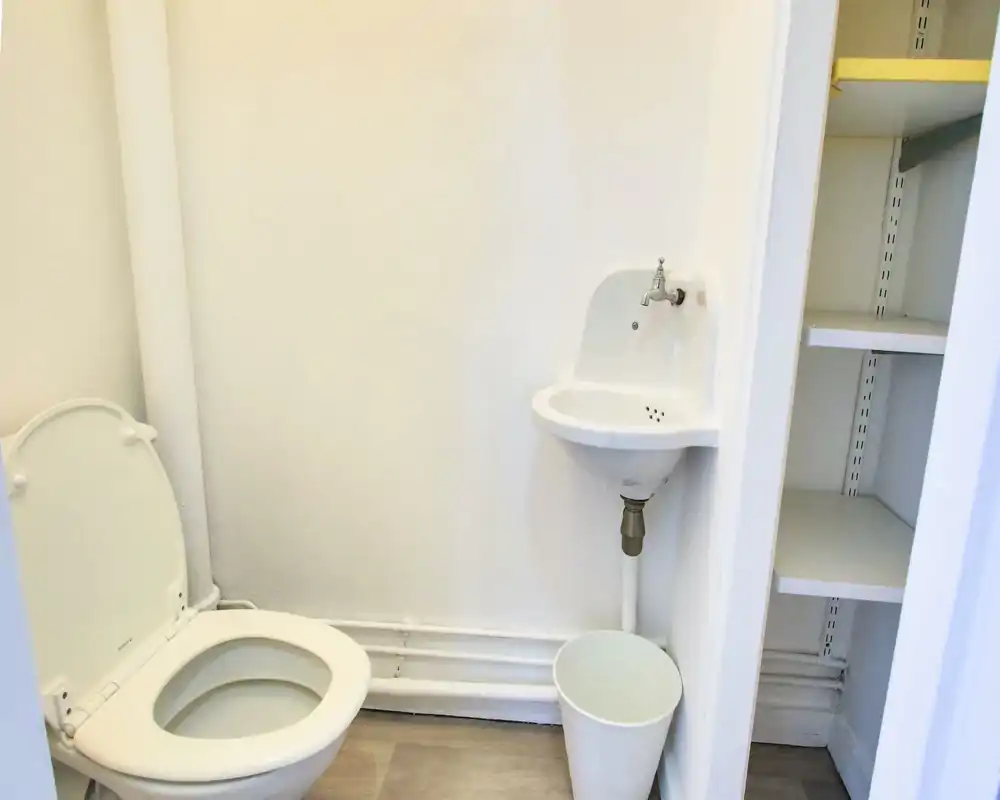 Small, bright bathroom with a toilet, a compact wall-mounted sink and built-in shelving; clean white finishes and light flooring create a neat, functional space.