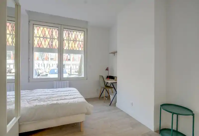 Camera da letto luminosa e minimal con letto bianco sotto una grande finestra e una piccola scrivania — ideale per una persona o uno studente.