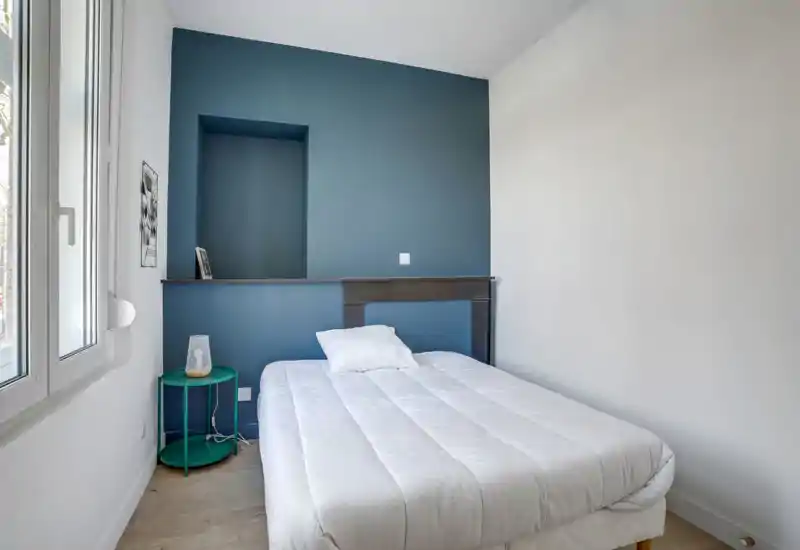 Dormitorio luminoso y minimalista con cama doble frente a una pared azul, luz natural de una gran ventana y una mesita de noche con lámpara.