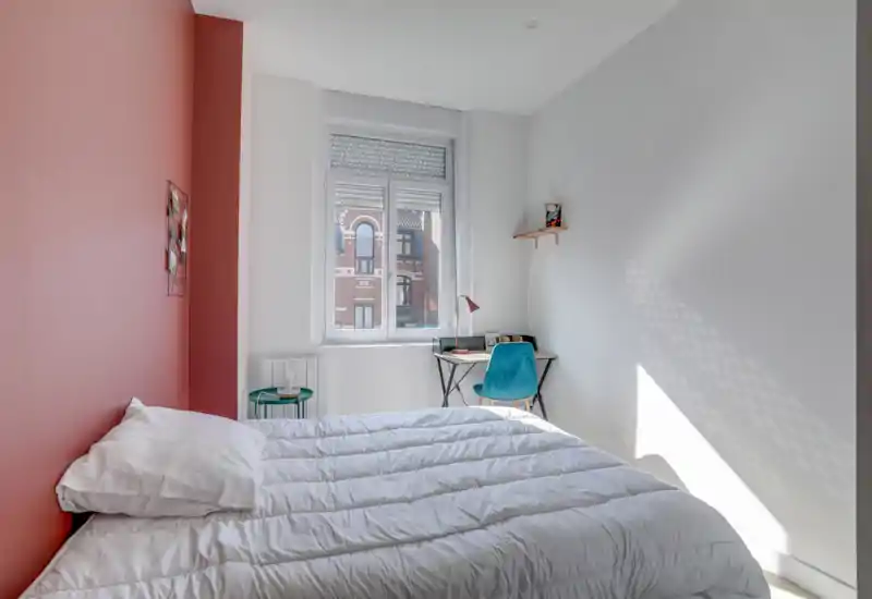 Camera da letto luminosa e ordinata con piumone bianco e parete d'accento color corallo. La luce naturale entra da una grande finestra; una scrivania compatta e una sedia blu creano un piccolo angolo studio.