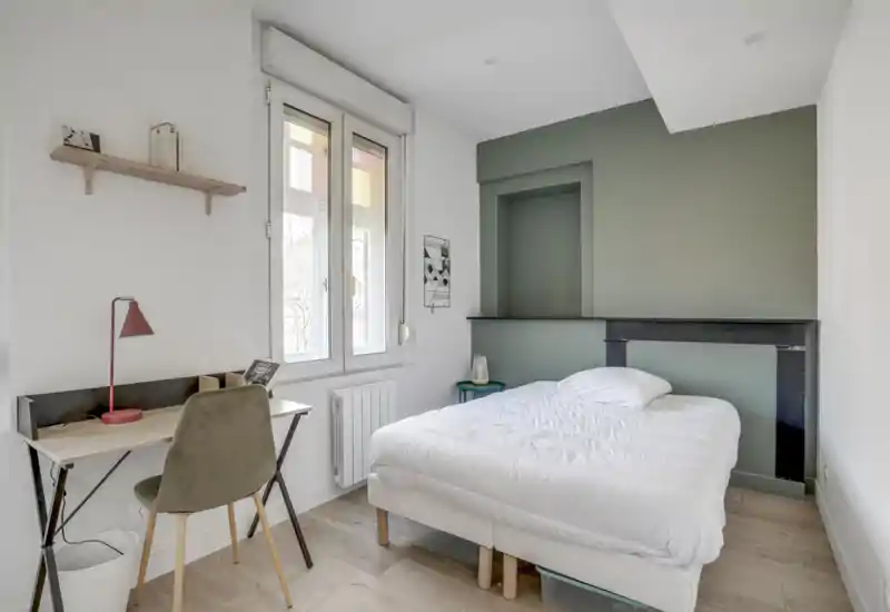 Chambre lumineuse et minimaliste avec un lit double bien fait contre un mur vert, une petite table de chevet et un bureau près de la fenêtre offrant une belle lumière naturelle.