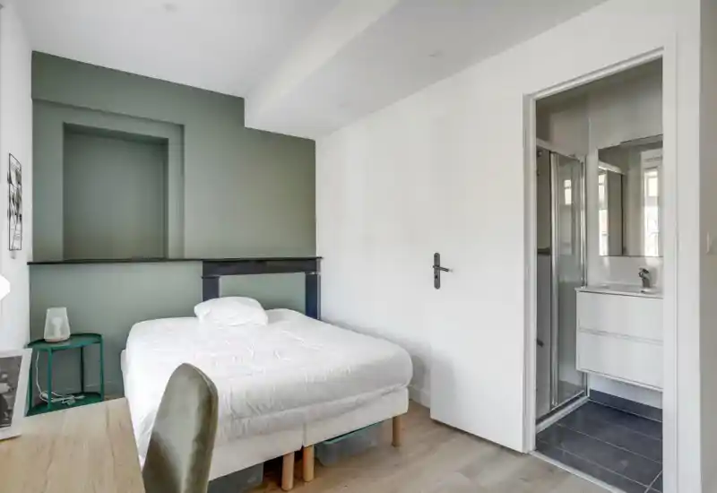 Chambre lumineuse et bien mise en scène avec un lit double devant un mur vert discret, une petite table de chevet et un accès direct à une salle d'eau compacte.