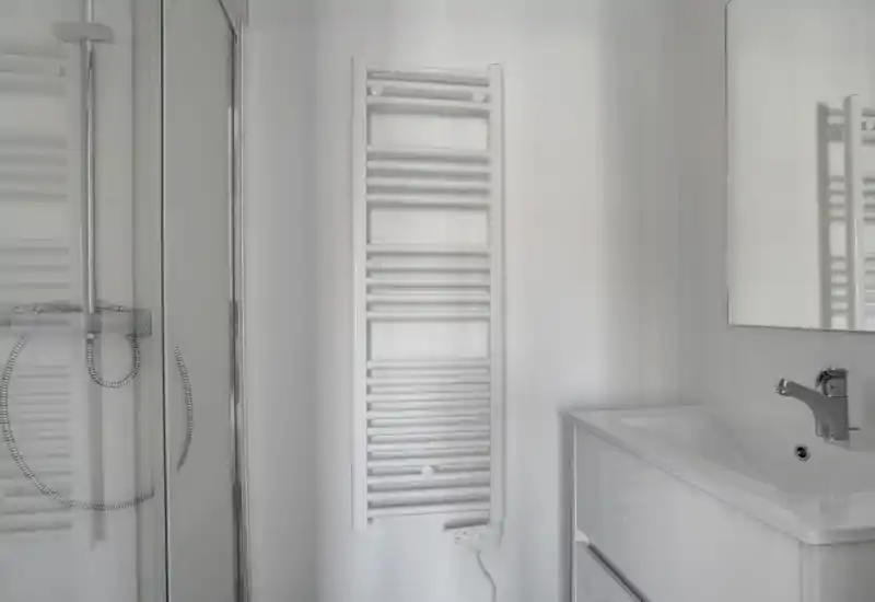 Salle de bain lumineuse et minimaliste avec douche en verre, radiateur porte-serviettes mural et meuble vasque blanc avec robinet.