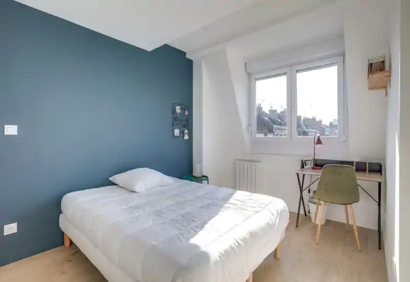 Dormitorio luminoso y bien arreglado con una cama doble cómoda junto a una pared azul y un pequeño escritorio junto a la ventana. La luz natural llena la habitación y el estilo moderno y sencillo crea un ambiente tranquilo.