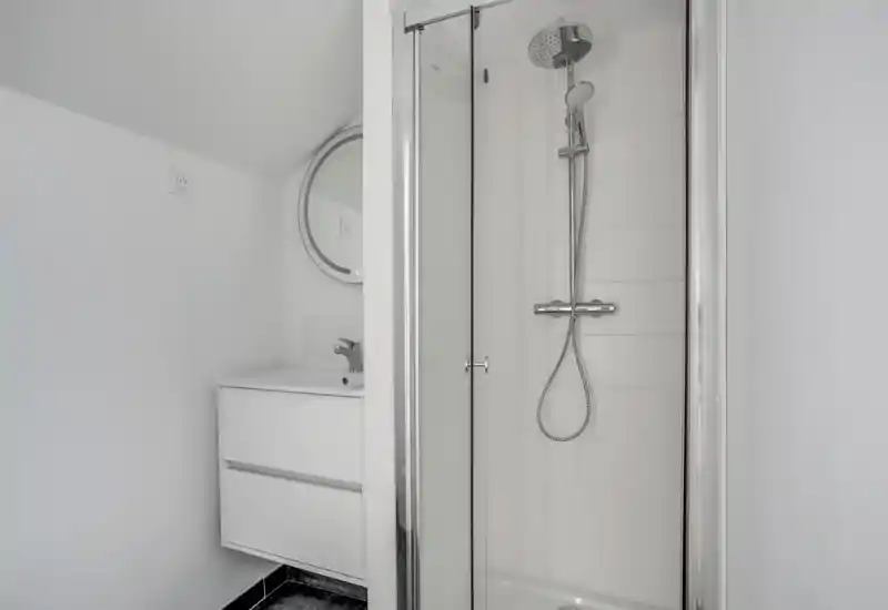 Baño moderno y minimalista con ducha acristalada, columna con rociador y teleducha, y un mueble lavabo compacto con espejo redondo. Azulejos blancos y grifería cromada dan una sensación luminosa y contemporánea.