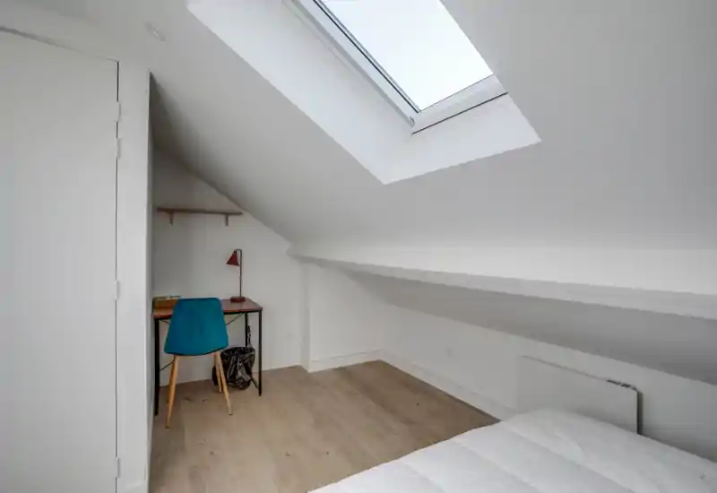 Angolo di camera mansardata luminoso con lucernario, vista parziale di un letto ben rifatto in primo piano e una piccola scrivania con sedia turchese e lampada — un angolo notte/lavoro compatto e minimal.