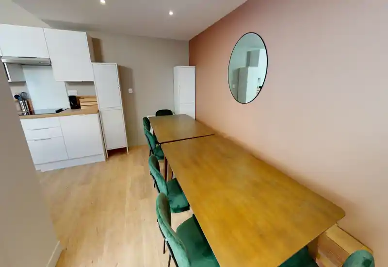 Spacious dining area with a long wooden table and green upholstered chairs, adjacent to a compact kitchenette. The soft peach wall and round mirror add a warm, contemporary touch.