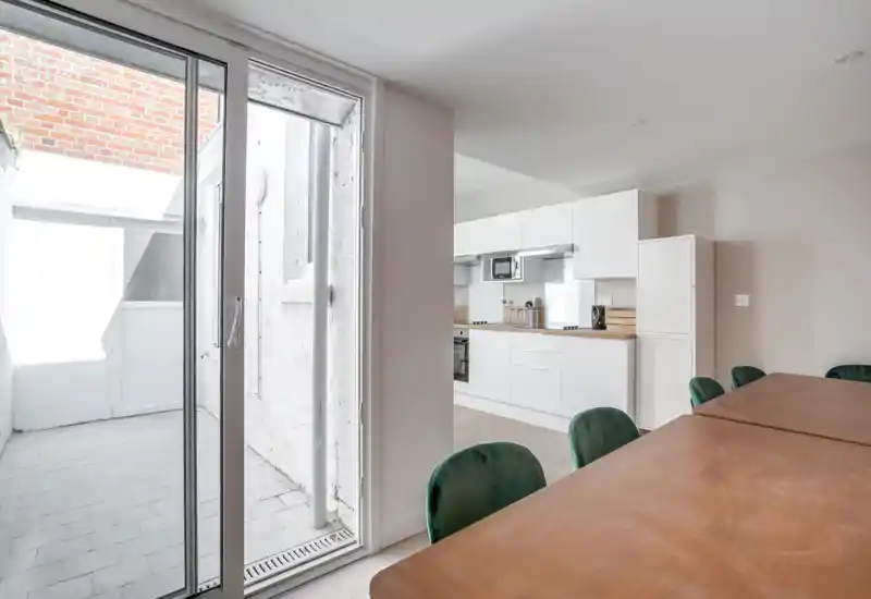Bright, modern open-plan kitchen with white cabinets and a wooden dining table with green upholstered chairs, plus a sliding glass door leading to a small outdoor area.