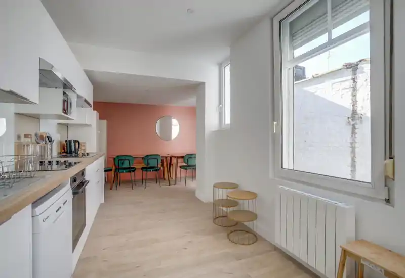 Cocina moderna y luminosa en concepto abierto con armarios blancos, encimera de madera y una pequeña zona de comedor frente a una pared terracota. Ventana grande que aporta luz natural, sillas verdes y decoración minimalista.