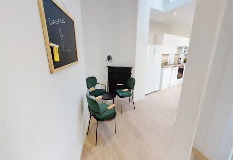 Pequeña zona de estar con cuatro sillas verdes tapizadas, una mesa redonda pequeña y una lámpara de pie junto a una repisa negra decorativa; espacio luminoso que conecta con la cocina blanca.