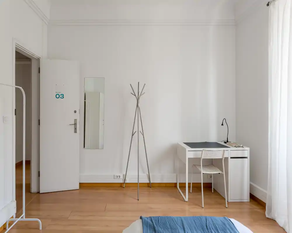Bright, minimal bedroom corner with a small desk, coat stand and a partially visible bed in the foreground. Clean white walls and natural wood flooring create a calm, airy atmosphere.