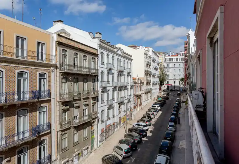 View of a sunny urban street from a balcony, showing parked cars and classic multi-story buildings with decorative facades under a blue sky.