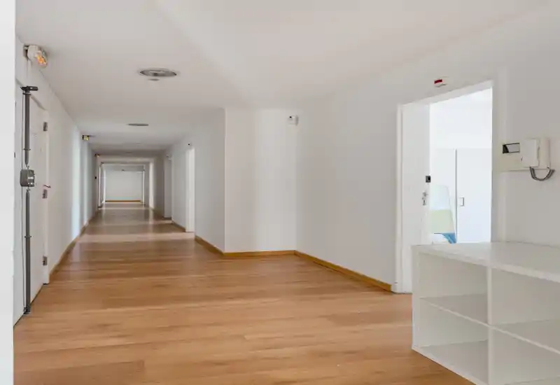 Bright, long hallway with wooden floors and white walls, suitable for an entrance or common corridor. Clean, minimal design with natural light at the far end.