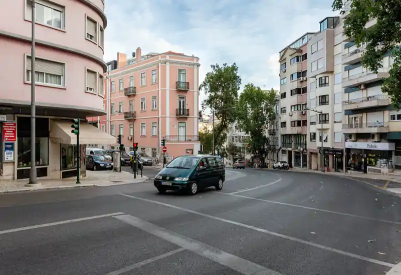 Street-view outdoor scene showing a curved urban intersection with multi-storey buildings, a parked car and light traffic — bright, clear and suitable to advertise proximity to local amenities.