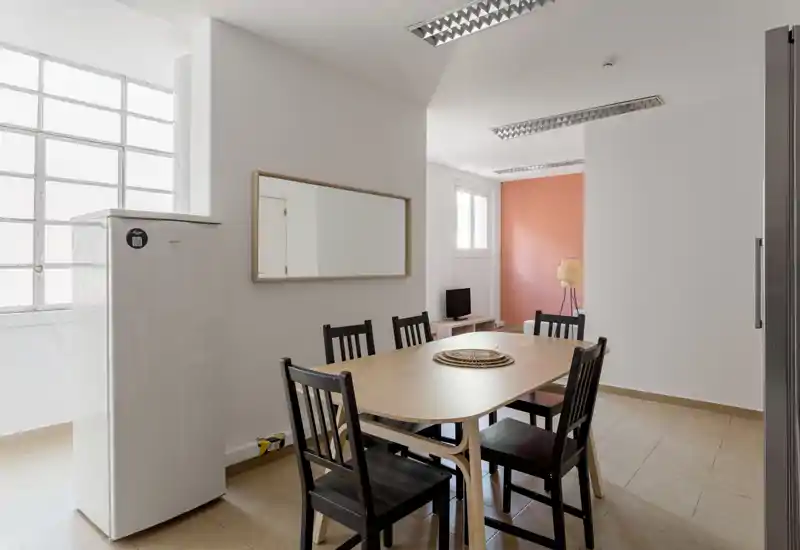 Bright, minimalist dining area with a wooden table and six chairs, an accent wall and a nearby fridge — ideal for small-group meals and casual gatherings.